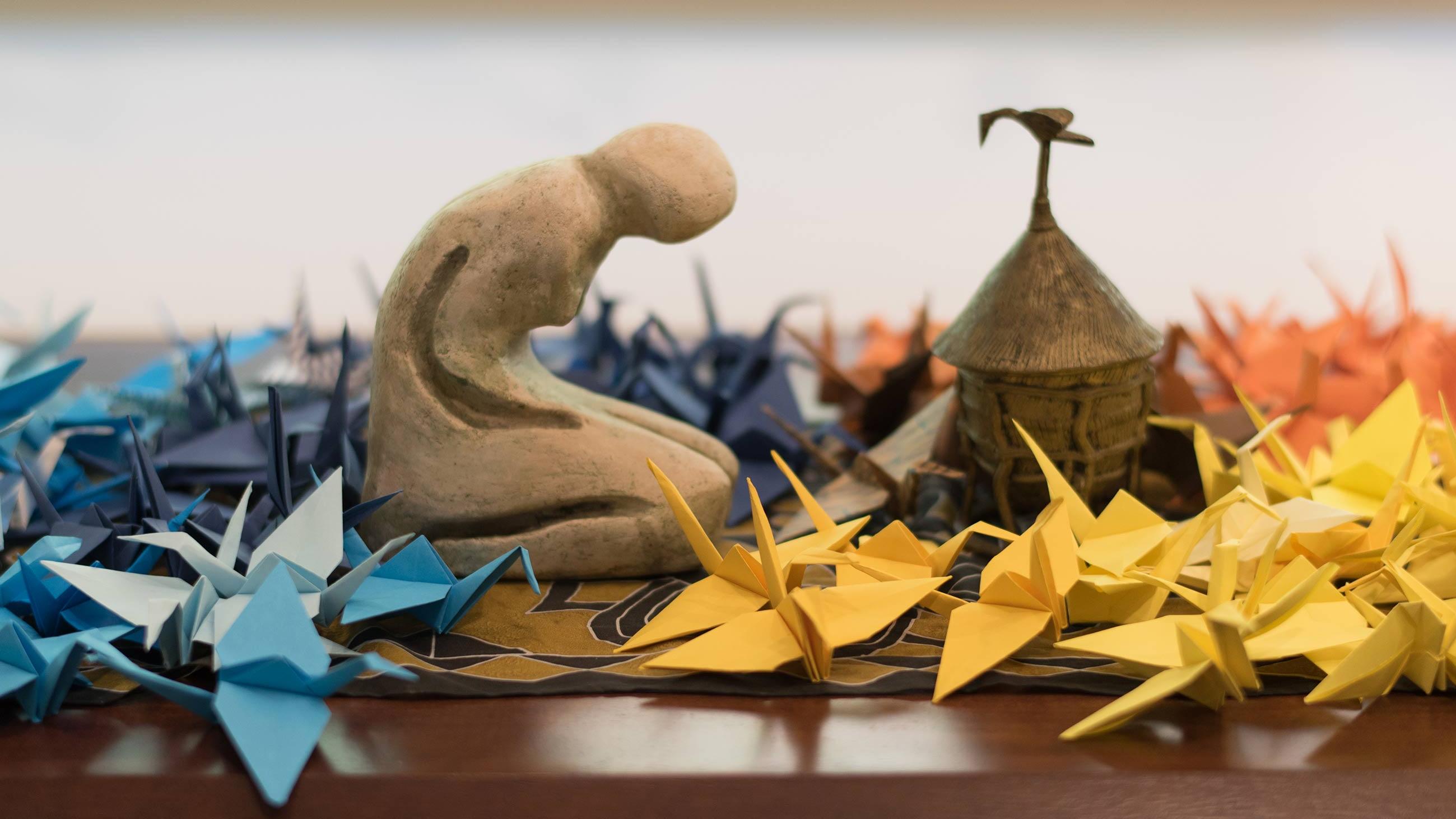 Small statue kneeling among multi-colored paper cranes